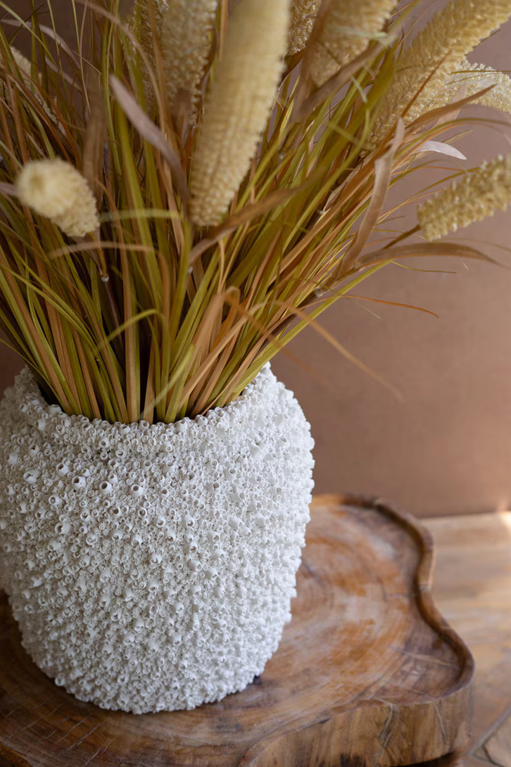 Decorative vase with textured surface holding a arrangement of grasses and plants on a wooden surface.