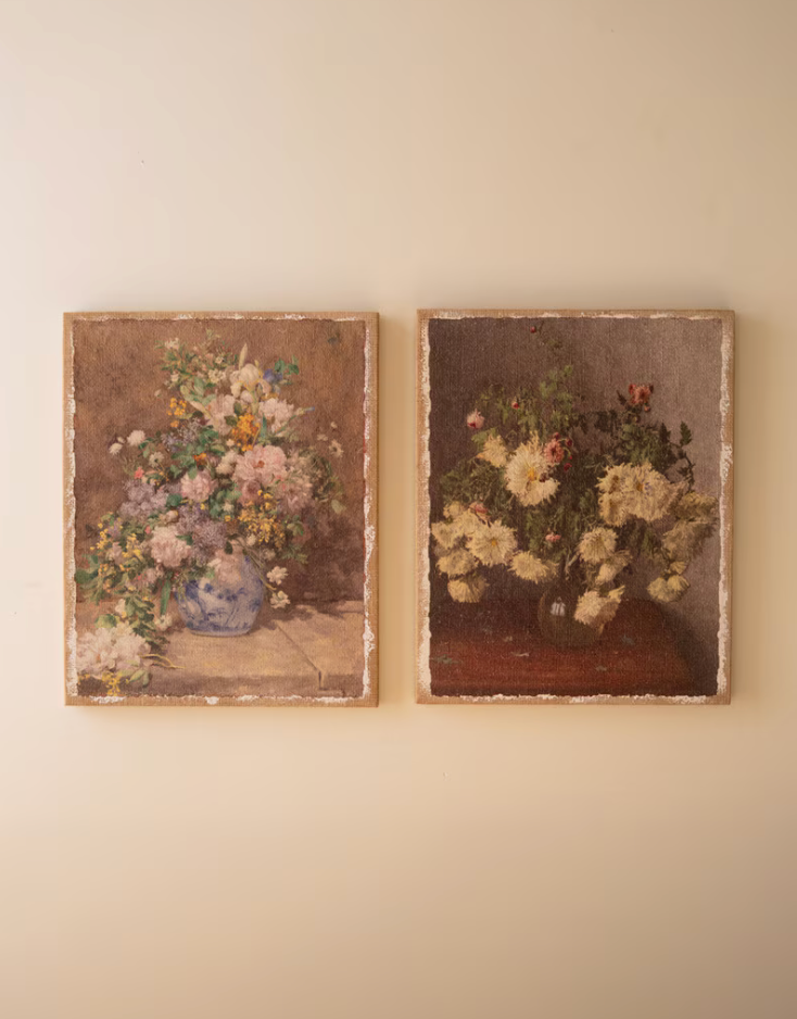 Two vintage framed oil paintings of floral still lifes hanging side-by-side on a warm beige wall. The left painting features a vibrant bouquet of pink, white, and yellow flowers in a blue-and-white vase against a brown background.
