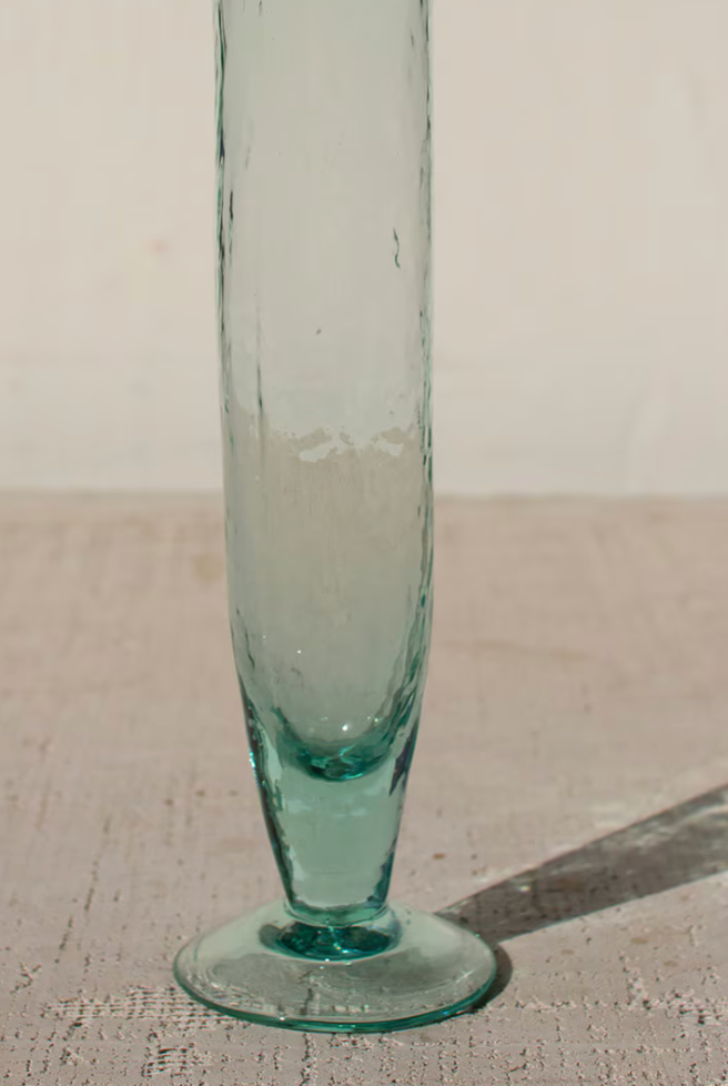 A tall, slender light blue-green glass vase with a flared base and circular foot, standing on a textured beige surface against a plain off-white background.
