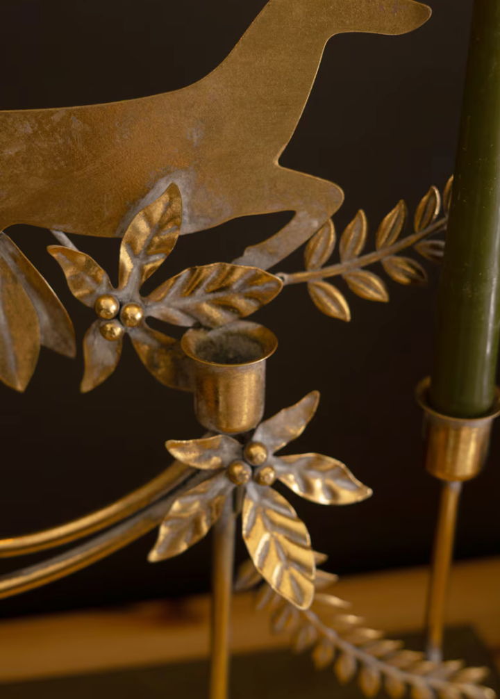 Close-up of a brass holiday wreath candle holder with leaf and pine branch details, holding a green candle, against a dark background.