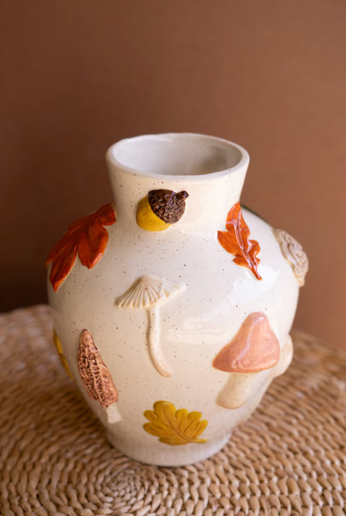A speckled cream ceramic vase decorated with raised autumn-themed motifs including mushrooms, acorns, and leaves in orange, yellow, brown, and pink, displayed on a woven placemat against a warm brown background.