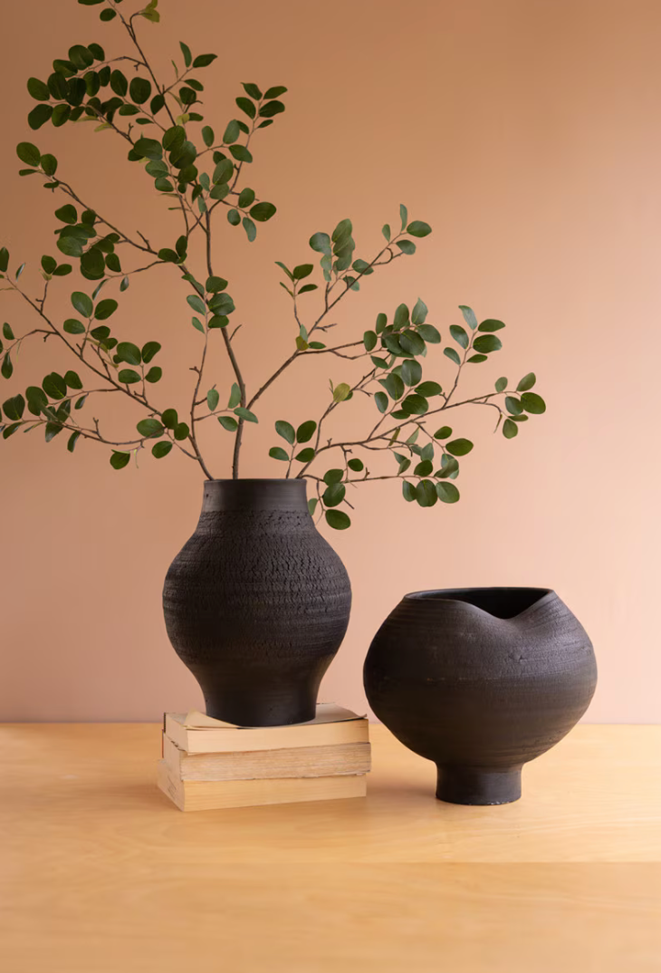 Two matte black ceramic vases on a light wood surface against a warm peach background. One vase stands on stacked books and holds leafy green branches, while the other is an open, rounded vessel beside it.