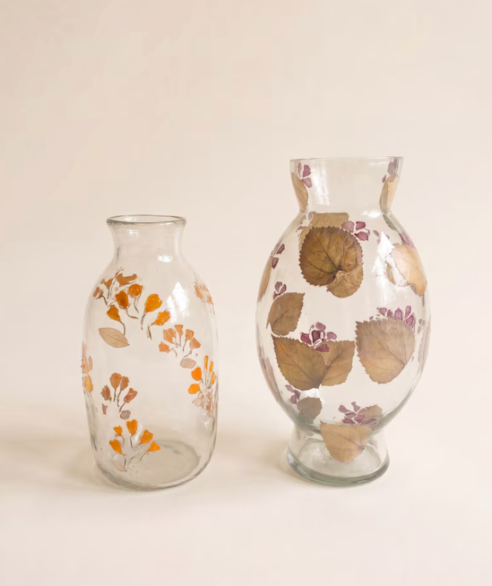 Two clear glass vases decorated with dried botanical elements, one small and one larger, standing on a light beige background.