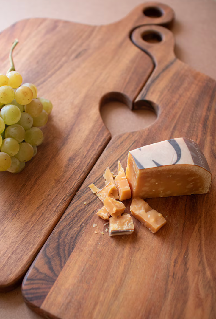 A heart-shaped wooden serving board with a handle, styled with a wedge and chunks of marbled cheese and a bunch of green grapes on a neutral background.