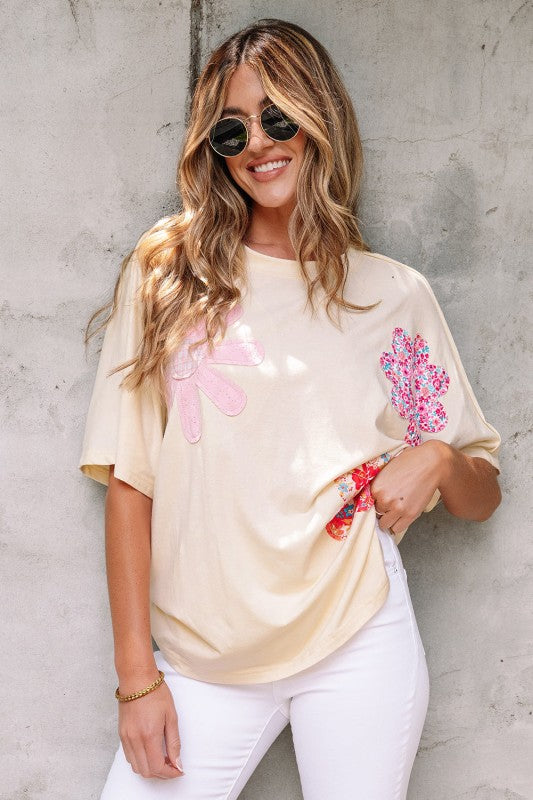 Woman wearing a beige t-shirt with floral designs and white pants against a concrete wall.