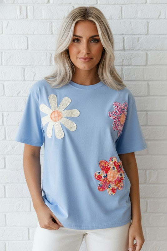 Woman wearing a light blue t-shirt with floral designs against a white brick wall.