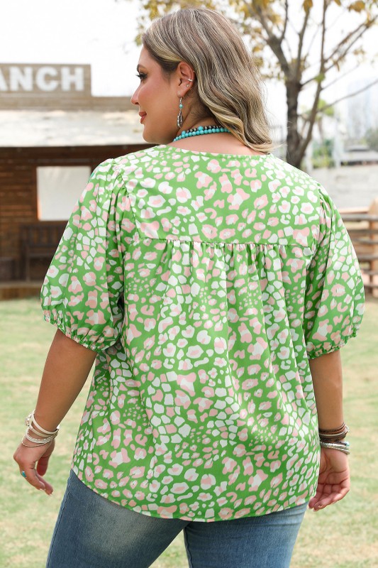 Woman wearing a green floral blouse outdoors with a wooden structure and trees in the background.