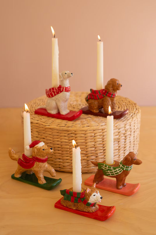 A set of dog-shaped candle holders with lit white candles, styled on a light wood table in front of a woven basket. The dogs are dressed in red, green, and plaid holiday sweaters and wear sled bases.