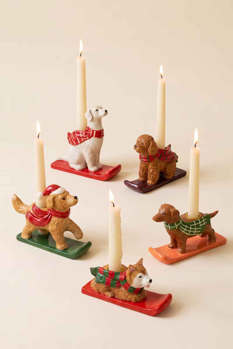 Five dog-shaped candle holders with lit taper candles, each wearing a different colored scarf and sitting on a sled base, arranged on a warm cream background.