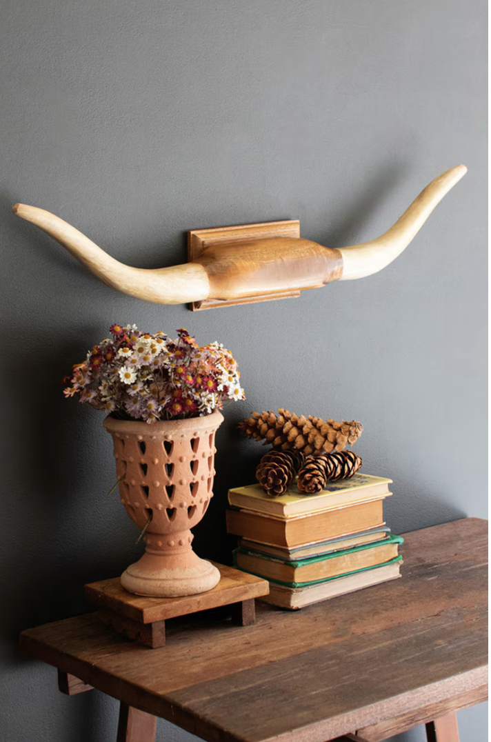 Decorative wall shelf with long horns, vase with flowers, and books on a wooden table against a gray wall.