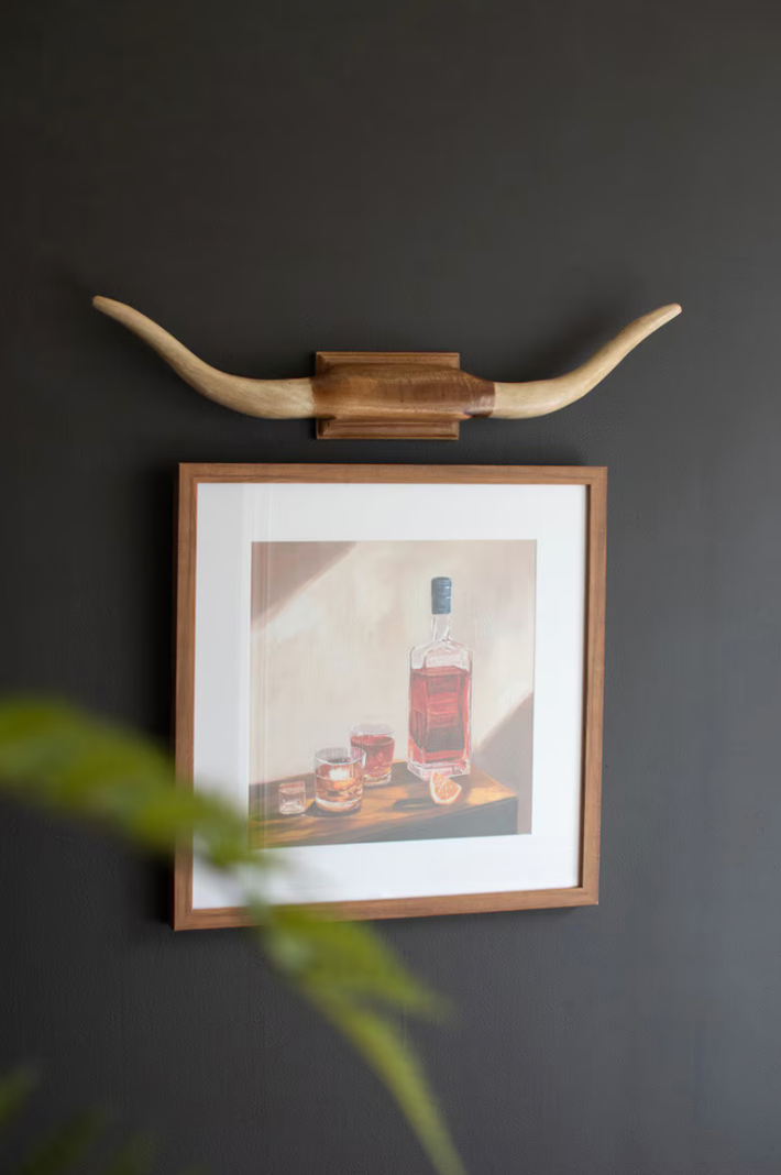 Framed artwork of a bottle and glasses on a dark wall with long horns above.