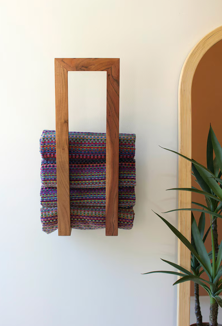 Wooden towel rack with folded multicolored towels against a white wall, with a plant in the corner.