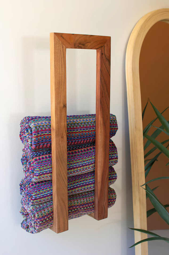 Stack of colorful folded blankets against a wooden frame on a white wall with a mirror and plant in the background.