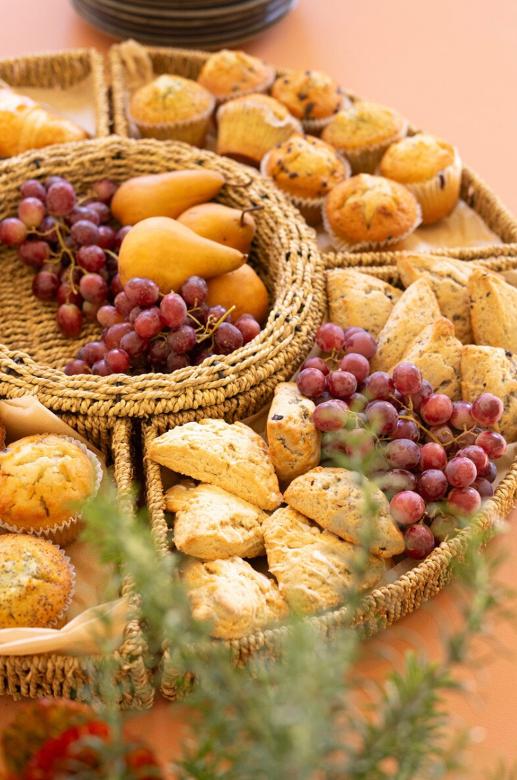 Assorted baked goods including muffins, scones, and grapes in a woven basket on a wooden table.