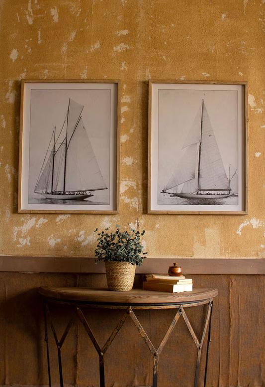 Two framed black and white sailboat prints on a textured yellow wall with a wooden table and plant below.