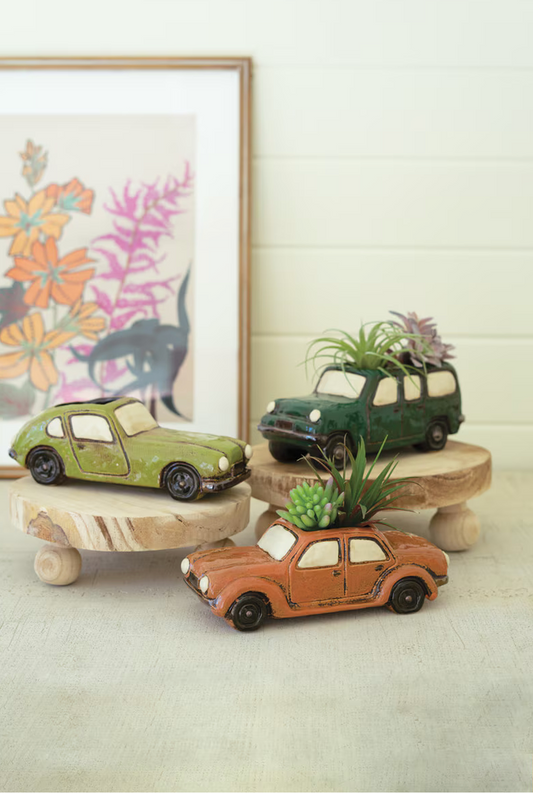 Three wooden car-shaped planters with plants on a light surface, framed artwork in the background.