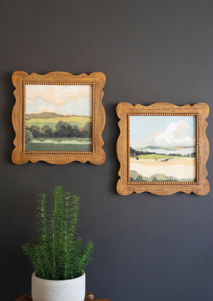 Two framed landscape paintings on a dark wall with a potted plant below.