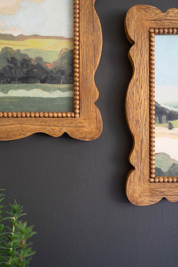 Two wooden-framed paintings on a dark wall with a plant in the foreground.