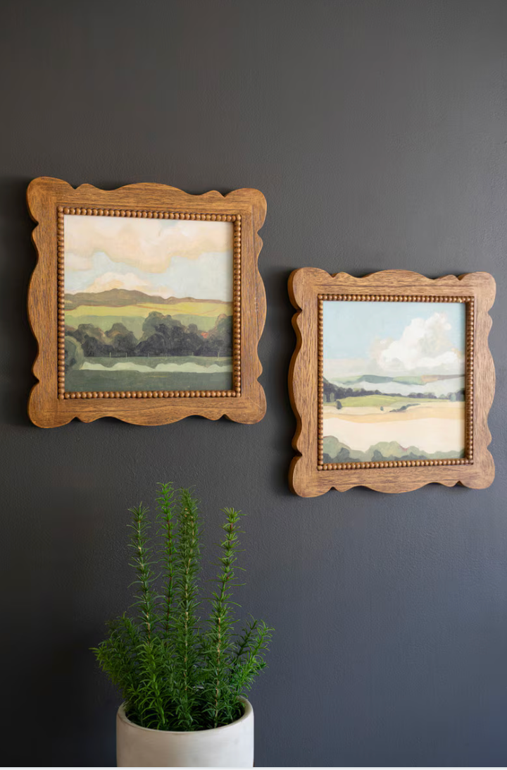 Two framed landscape paintings on a dark wall with a potted plant in the foreground.