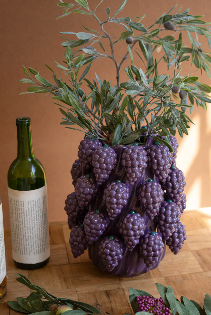 Purple decorative grapes in a vase on a wooden surface with a wine bottle in the background.