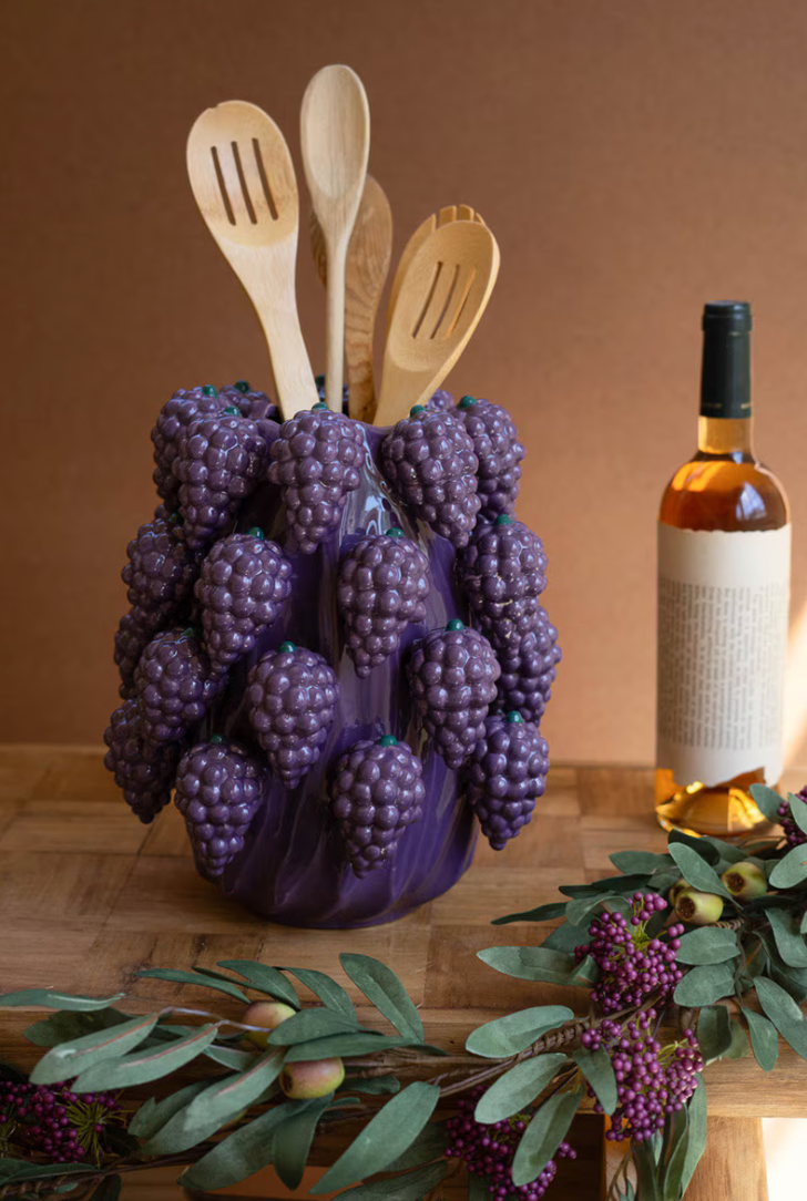 Grape-shaped utensil holder with wooden spoons on a wooden surface with a bottle of wine in the background.