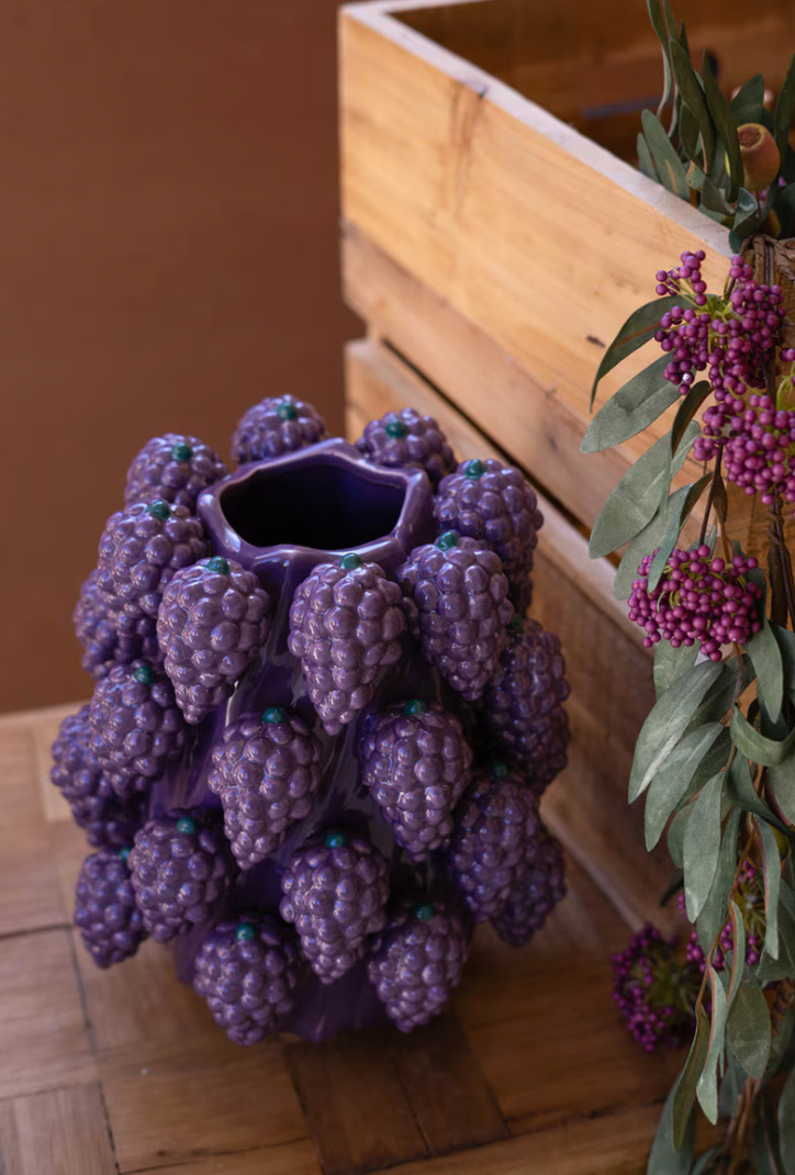 Vase shaped like a bunch of purple grapes on a wooden surface with a plant in the background.