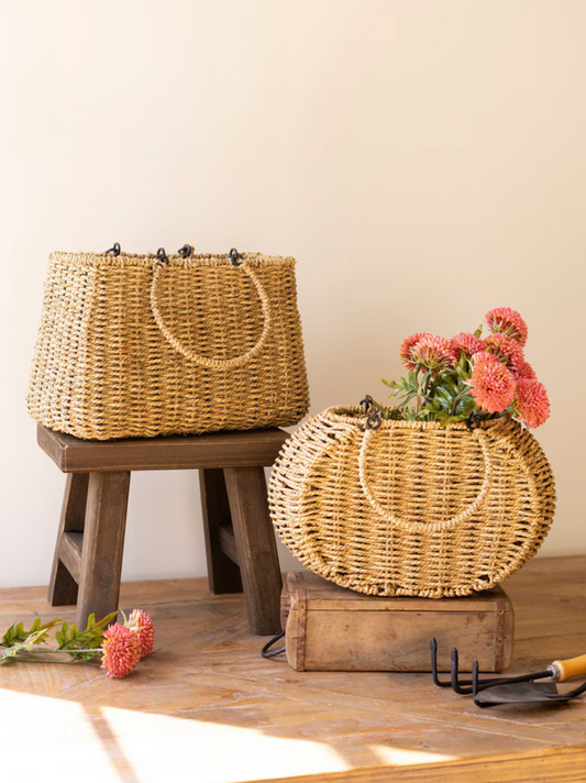 two woven baskets that present like purses with flowers inside and placed on a table with garden tools
