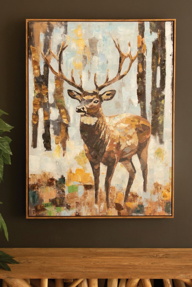 Framed painting of a deer in a forest setting on a wall.