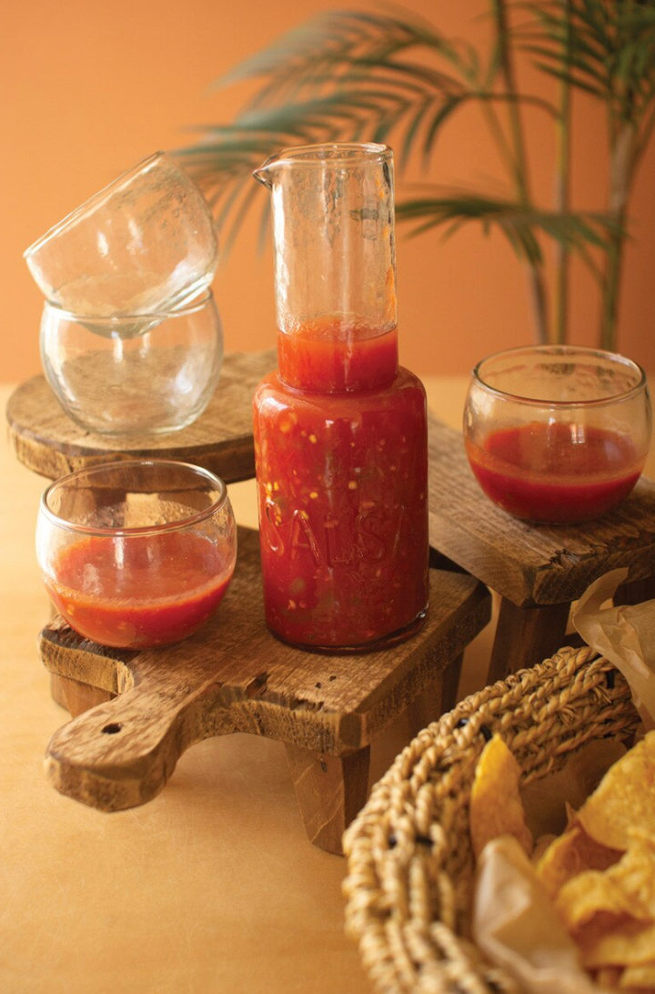 jar of salsa and 2 empty bowls and 2 bowls filled with red salsa on a wooden stool with an orange background and chips on the side in a bowl
