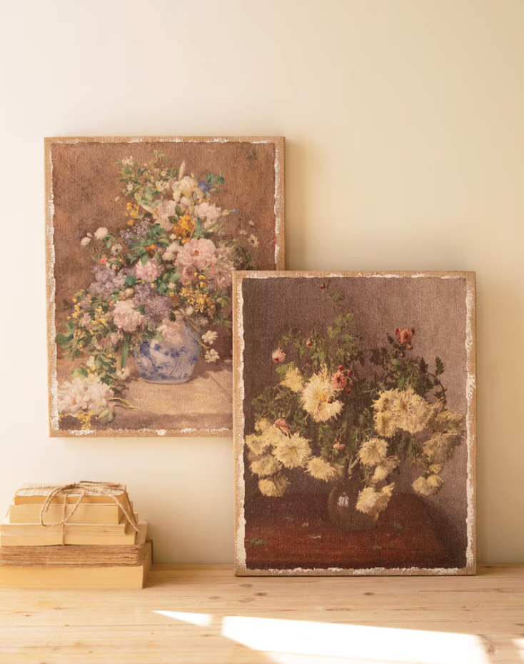 Two framed vintage-style floral paintings leaning against a cream wall on a light wood floor, with a small stack of books tied with twine in the foreground.