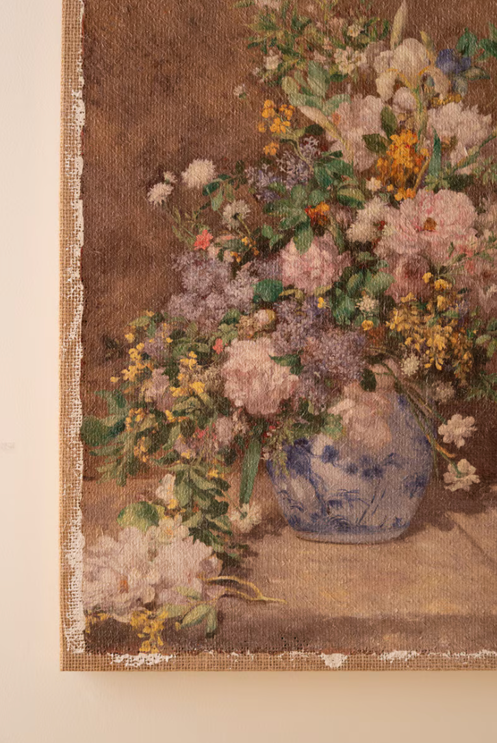 A close-up of a floral still life artwork or tapestry showing a blue-and-white vase filled with pink, cream, yellow, and lavender flowers against a warm brown background, mounted on a light beige wall.
