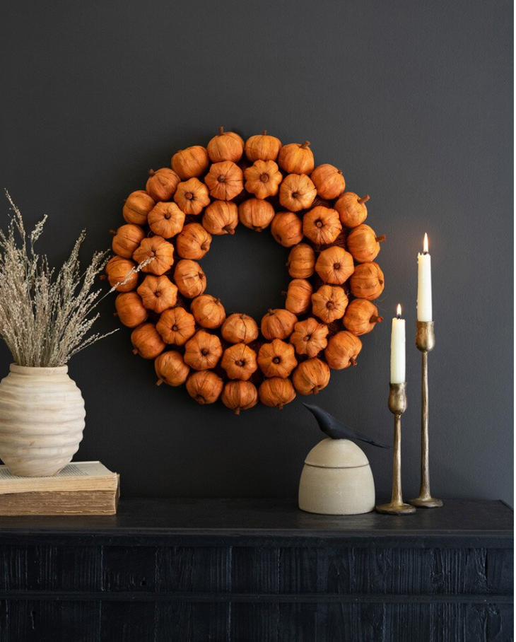 A circular autumn wreath made of small orange pumpkins hangs on a dark charcoal wall above a mantel styled with a cream vase of dried grass, books, a ceramic bird figurine, and two lit candlesticks.