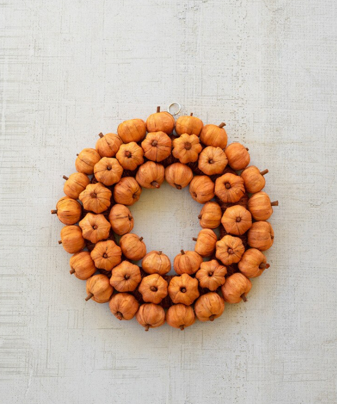 A circular autumn wreath made of small orange-brown pumpkins arranged in a ring, with a silver hanging loop at the top, photographed against a light textured background.