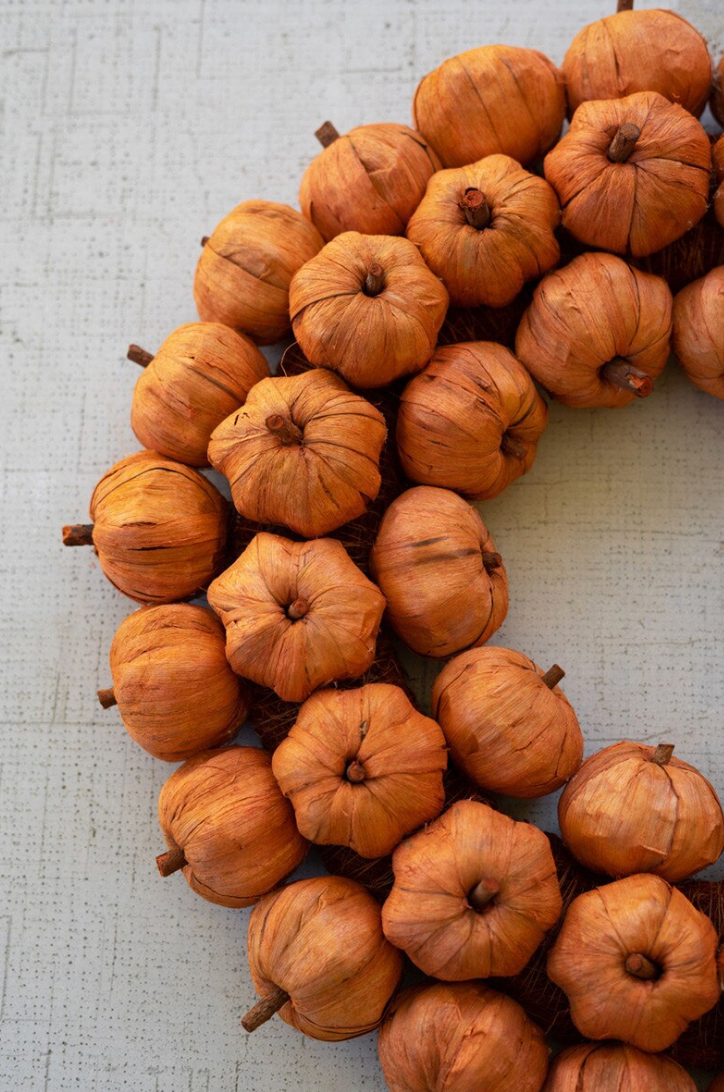 A decorative wreath made of small dried orange-brown pumpkins arranged in a curved shape on a light textured fabric background.
