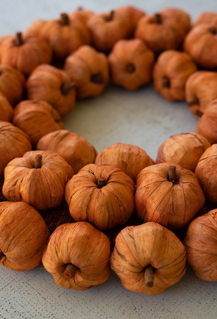 A circular autumn wreath made of small dried pumpkins or gourds, arranged in a ring on a light neutral surface.