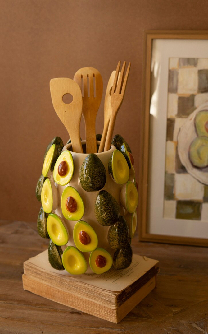 A ceramic kitchen utensil holder decorated with sliced avocado pieces, holding wooden spoons and forks, set on a stack of books against a warm brown wall.