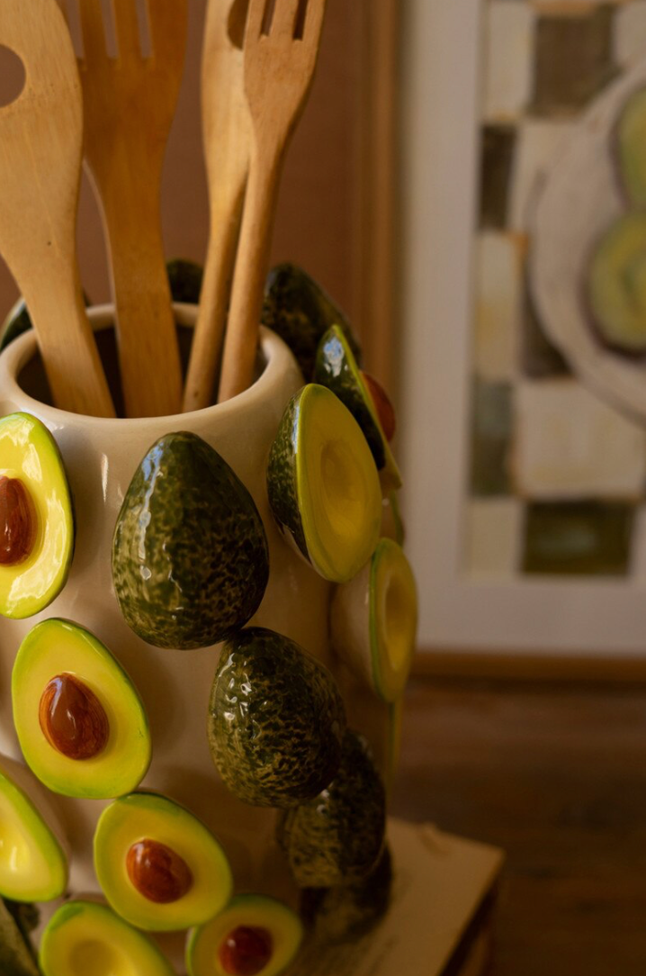 A ceramic kitchen utensil holder decorated with 3D avocado shapes and avocado slice patterns, filled with wooden spoons and forks, in a warm indoor setting.