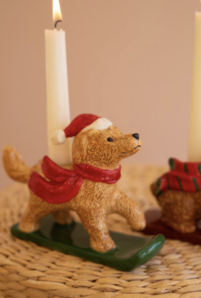 A ceramic dog-shaped candle holder wearing a red Santa scarf and red-and-white Santa hat, standing on a green sled base with a lit white candle inserted. The item is styled on a woven placemat with a softly blurred warm beige background.