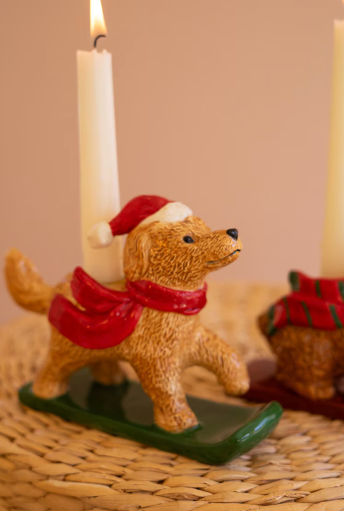 A ceramic dog-shaped candle holder wearing a red Santa scarf and red-and-white Santa hat, standing on a green sled base with a lit white candle inserted. The item is styled on a woven placemat with a softly blurred warm beige background.