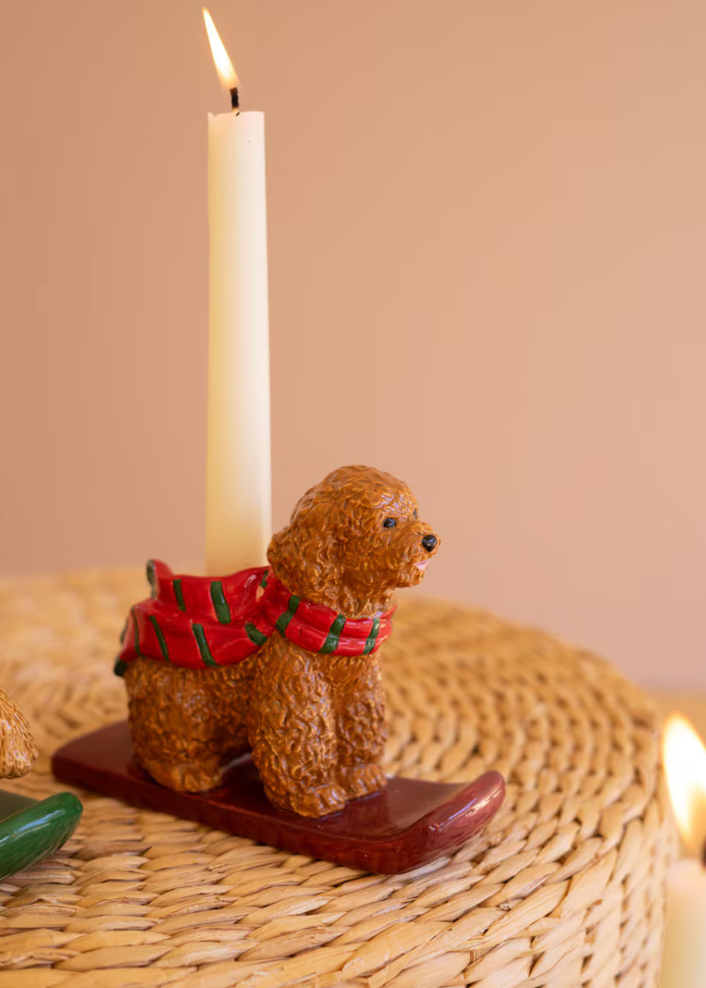 A small brown poodle figurine wearing a red and green plaid scarf, mounted on a dark wooden base and holding a lit white candle. The scene is set on a woven round mat against a soft beige background.