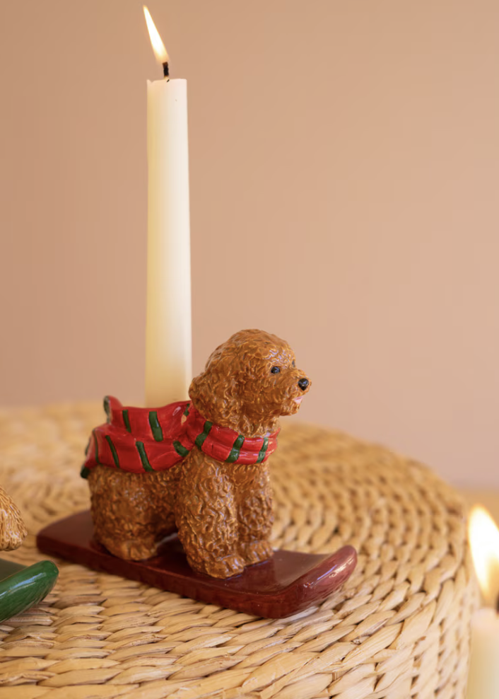 A small brown poodle figurine wearing a red and green plaid scarf, mounted on a dark wooden base and holding a lit white candle. The scene is set on a woven round mat against a soft beige background.