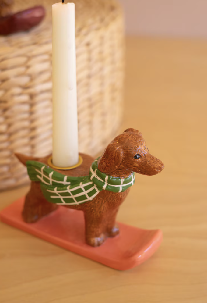 A small brown dog-shaped candle holder on a coral-orange base, wearing a green plaid scarf, holding a white taper candle on a wooden tabletop with a blurred woven basket in the background.