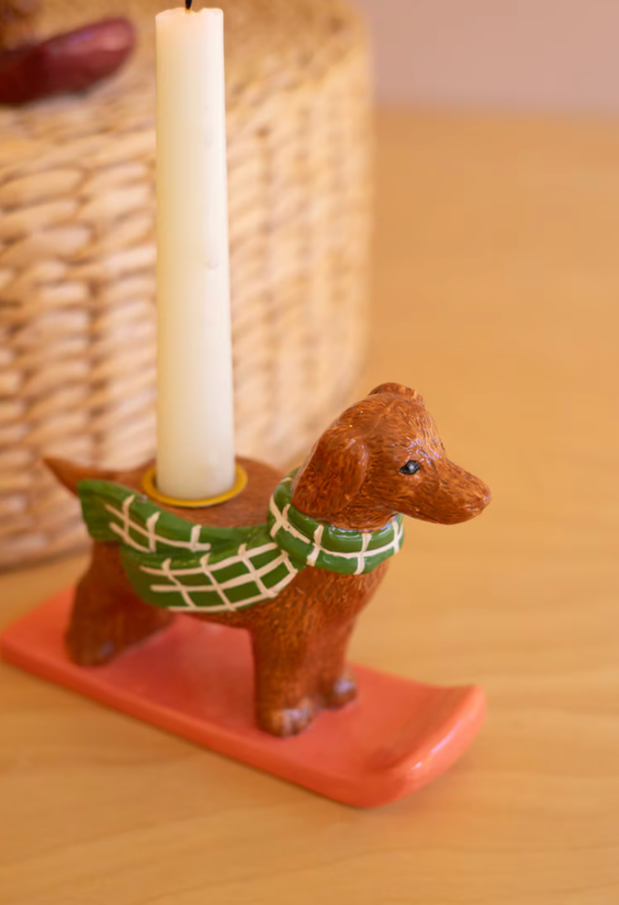 A small brown dog-shaped candle holder on a coral-orange base, wearing a green plaid scarf, holding a white taper candle on a wooden tabletop with a blurred woven basket in the background.