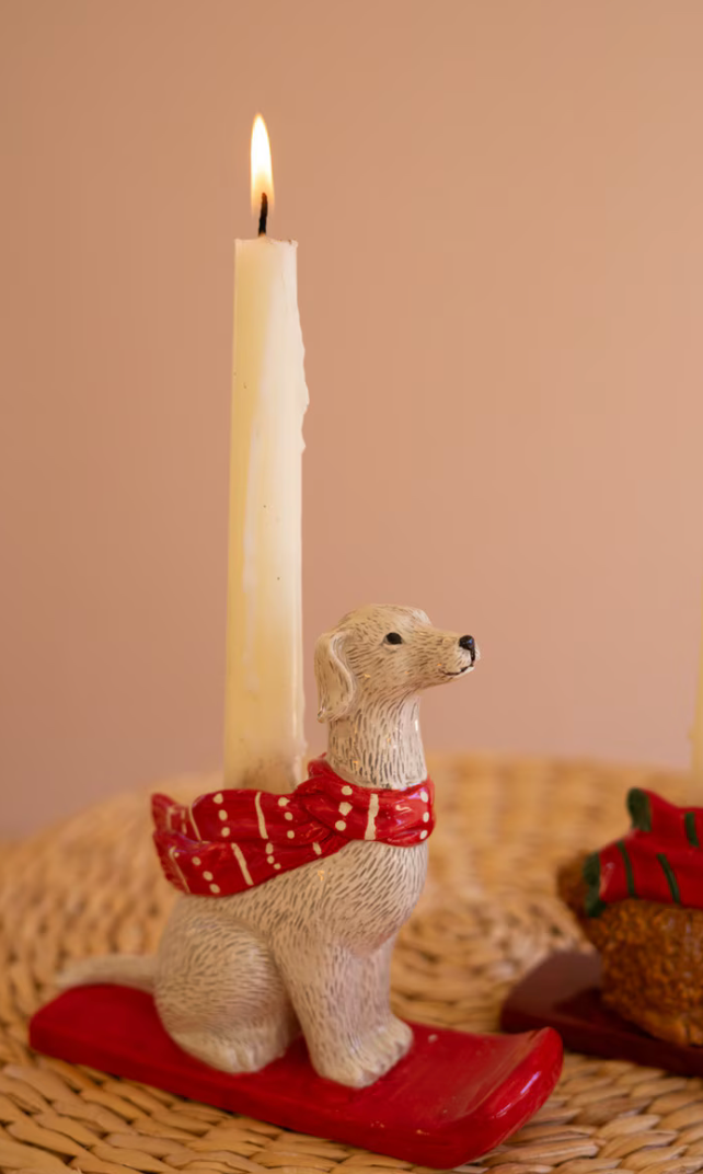 A lit white candle in a dog-shaped holder wearing a red patterned scarf and sitting on a red sled, placed on a woven surface against a warm beige background.