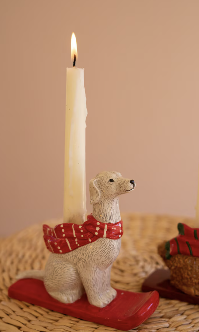 A lit white candle in a dog-shaped holder wearing a red patterned scarf and sitting on a red sled, placed on a woven surface against a warm beige background.