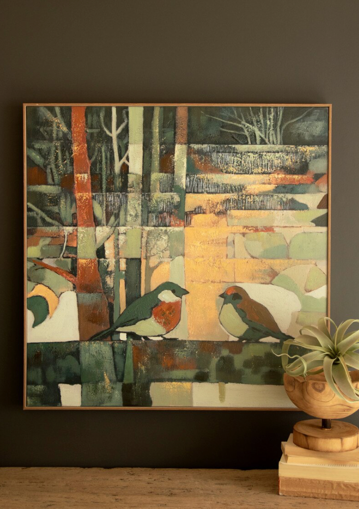A framed abstract bird painting hangs on a dark wall above a wooden surface, with a potted plant and books beside it. The artwork features two birds in a patchwork-style scene with muted greens, beige, orange, gray, and white tones.