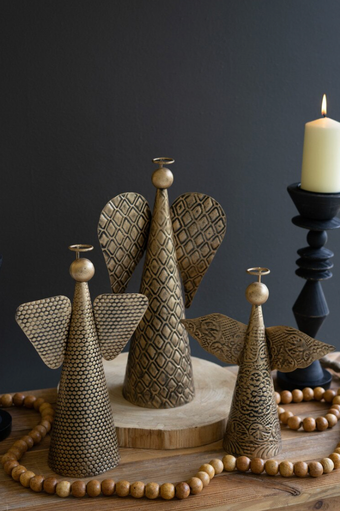 Three decorative angel figurines with textured wings and cone-shaped bodies in a metallic bronze finish, arranged on a wooden surface with a bead garland and a lit candle in the background against a dark gray wall.