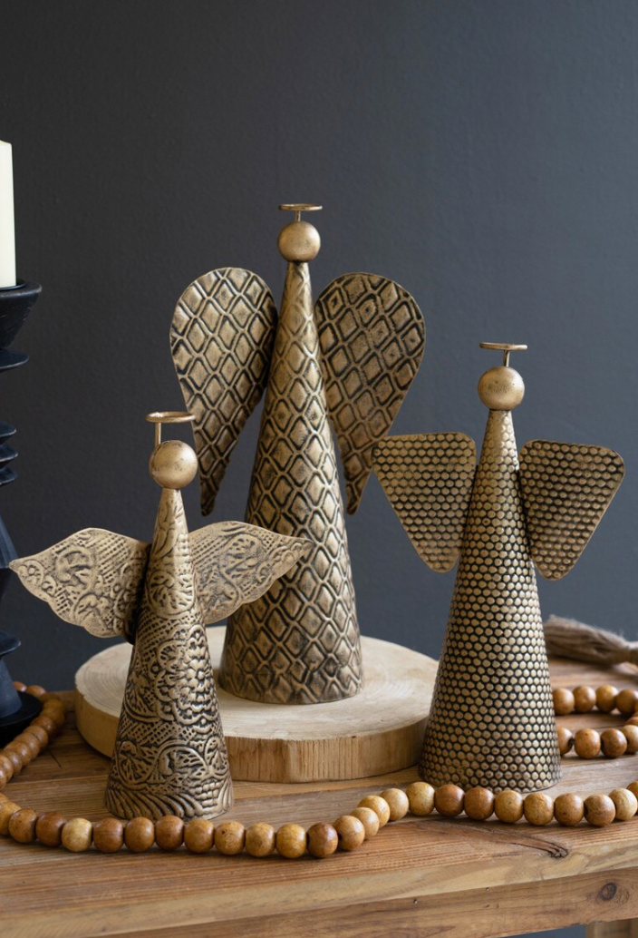 Three decorative angel-shaped metal ornaments with textured wings and cone bodies, arranged on a wooden surface against a dark gray background.