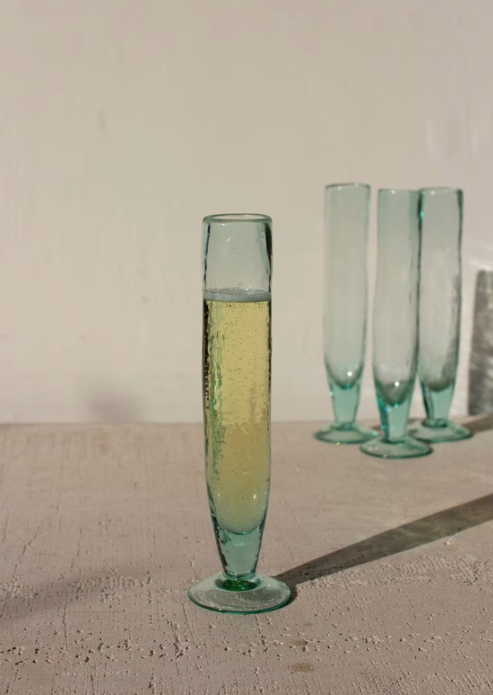 A set of translucent light blue-green glass flutes, with one in the foreground filled with pale yellow bubbly liquid and three more standing empty in the background against a plain off-white wall.