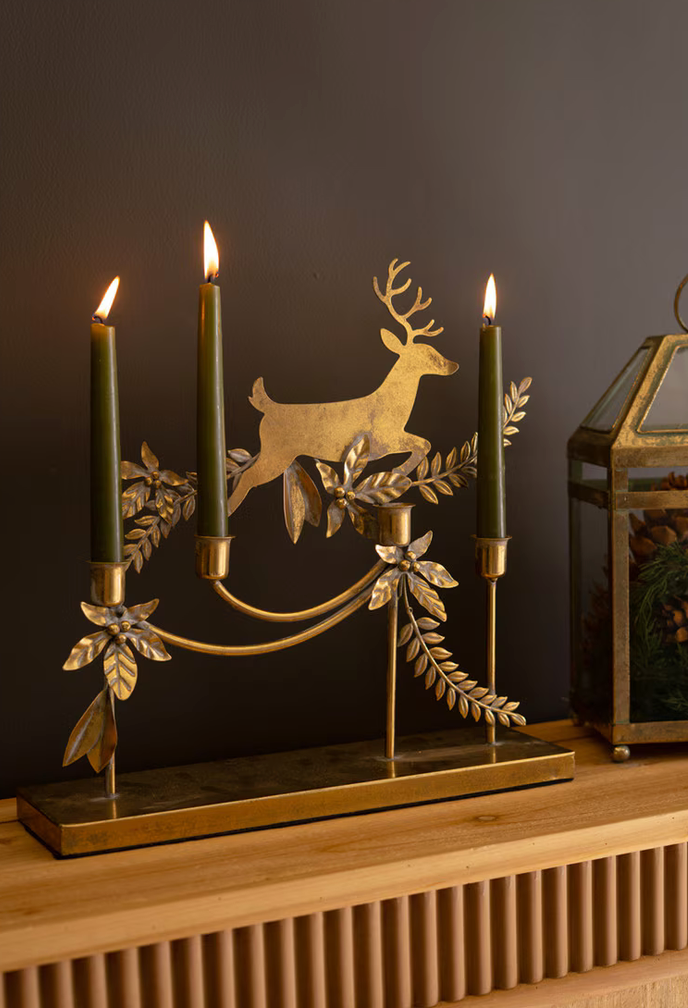 A brass candelabra with a deer silhouette and leafy branches, holding three lit dark green candles, displayed on a wooden mantel against a dark gray wall.